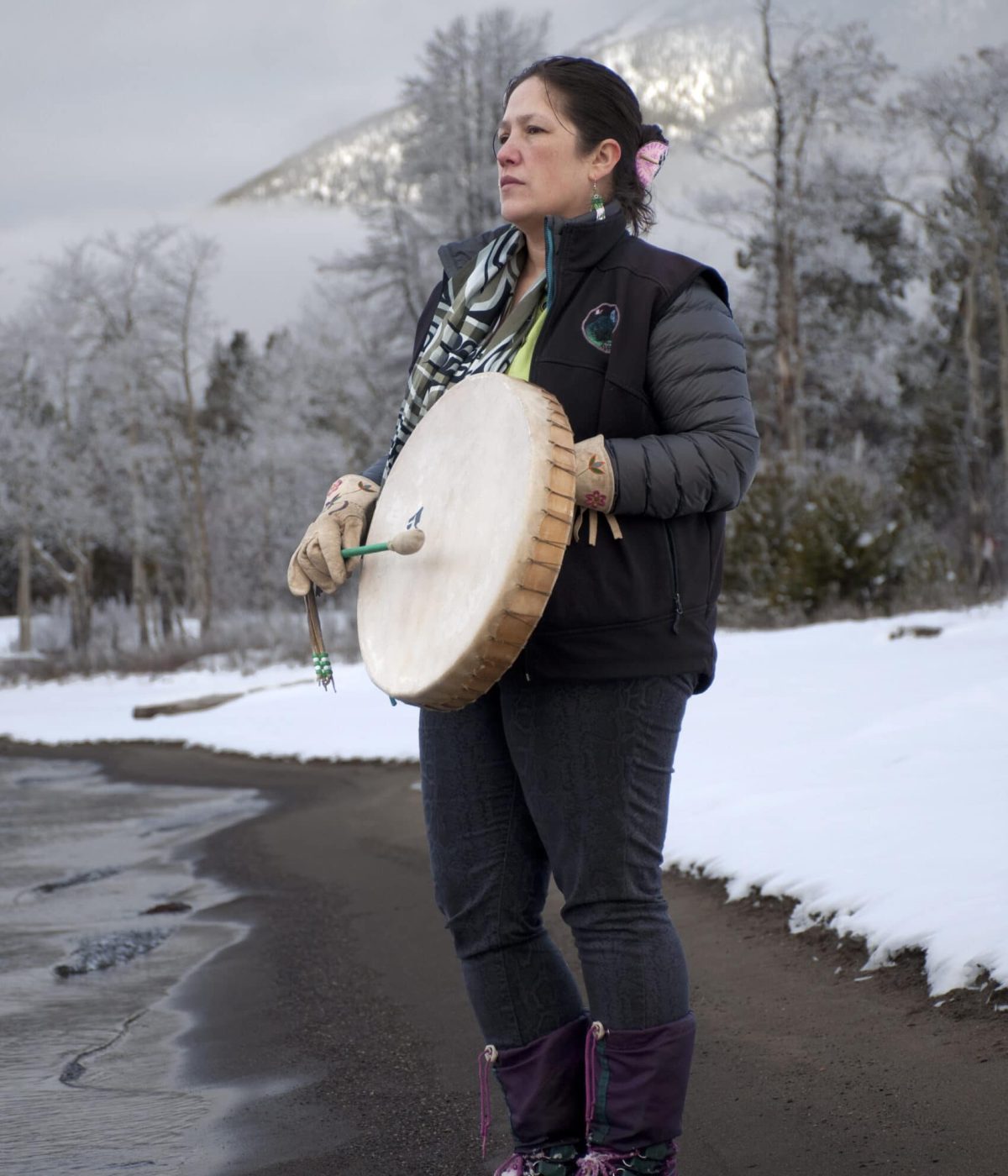 Marilyn with her drum at the edge of Chilko Lake with one of the many mountains in Xeni, Nemiah Valley, behind her.